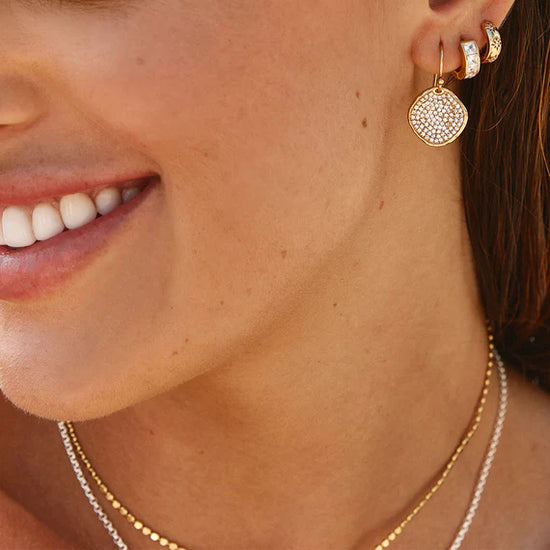 A close-up of a person wearing gold-colored round hook earrings with white round Swarovski crystals.
